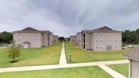 The balcony view from this space reveals a thoughtfully designed residential community characterized by its strong sense of symmetry and open outdoor environment. Two parallel rows of multi-story residential buildings flank a central green corridor, creating a visually organized and expansive outdoor setting. Each building features a neutral tan and brown exterior with pitched rooflines, multiple windows, and individual balconies that contribute to the architectural consistency throughout the community.

The grounds between the buildings are well-maintained, with broad stretches of neatly trimmed green lawn running the length of the corridor. A concrete walkway bisects the space down the center, providing a clear and accessible pedestrian path that extends deep into the community. The pathway is flanked by additional sidewalks along the building fronts, enhancing connectivity throughout the grounds.

HVAC units are visible along the building bases, and utility signage is positioned along the central walkway. The view is open to a wide, overcast sky, giving the outdoor space a sense of expansiveness. Mature trees are visible in the background, providing a natural boundary and adding greenery to the overall landscape.