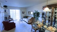 This single-floor residence offers a welcoming and functional layout centered around a combined living and dining area. The living space is anchored by a classic sofa with decorative fringe trim, complemented by a glass-top coffee table and plush wall-to-wall carpeting that adds warmth underfoot. A large sliding glass door floods the room with natural light and offers views of the surrounding greenery, creating a bright and airy atmosphere throughout the day. The dining area sits adjacent to the living space, featuring a glass-top dining table with ornate wooden chairs, elegantly set and ready for entertaining. Decorative touches throughout — including framed wall art, a tall indoor plant, and a traditional floor lamp — lend the space a classic, homey character. The open-plan arrangement between living and dining areas promotes a seamless flow, making the space feel generously proportioned and easy to navigate. The neutral wall tones and soft carpeting tie the rooms together cohesively, resulting in an interior that is both practical and comfortable.