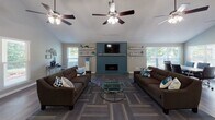 This well-appointed single-floor communal space offers an open, airy layout accentuated by dramatic vaulted ceilings and multiple ceiling fans that enhance airflow and comfort throughout. The central gathering area features rich, dark hardwood flooring that transitions to patterned carpet in the lounge zone, where two large brown sofas flank a glass-top coffee table, creating an inviting seating arrangement. A striking teal herringbone accent wall serves as the room's focal point, framing a fireplace with a stone surround and a mounted flat-screen television, flanked by floating shelves for display. Natural light streams in through multiple windows, offering views of lush greenery outside. A dedicated coffee bar station occupies one corner, while a built-in work counter with bar stools provides a functional workspace area. Tasteful wall art, including a triptych of abstract nature-inspired panels and a large decorative clock, adds character to the neutral gray walls. The kitchen area features gray shaker-style cabinetry, black appliances, a single-basin sink, and a wide peninsula with a chevron-paneled front and quartz-style countertop accommodating bar seating — ideal for casual dining or socializing. Recessed and flush-mount lighting ensures the space remains bright and welcoming throughout.