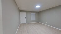 This well-maintained single-floor space offers a clean and modern aesthetic throughout, characterized by a consistent neutral gray color palette, crisp white trim, and light wood-look flooring that flows seamlessly across every room. The open layout provides a generous sense of space, with each area benefiting from flush-mount ceiling lighting and windows fitted with white horizontal blinds that allow for natural light control.

The main living area is bright and open, anchored by a front entry door and a window that introduces natural light. An adjacent open area with a stepped ceiling detail adds architectural interest while maintaining the space's cohesive design.

The kitchen is a standout feature, showcasing white shaker-style cabinetry with matte black hardware, a stone-look granite countertop, a black matte sink faucet, and a white tile backsplash. A stainless steel refrigerator and an overhead range hood complete the functional setup, with a peninsula counter providing additional workspace.

The bedroom area is quietly tucked away, offering a windowed wall with ample wall space and continued light flooring. The bathroom is elegantly appointed with large-format marble-style wall tiles surrounding the soaking tub and shower combination, complemented by matte black fixtures throughout, delivering a refined, contemporary finish.