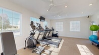Located at 5500 Hildebrand Rd. in Columbus, Ohio, this fitness center offers a compact yet fully functional workout space designed to support a range of exercise routines. The single-room facility is finished with neutral-toned carpet flooring and crisp white walls, creating a clean and inviting atmosphere throughout. Multiple windows fitted with horizontal blinds line the walls, allowing generous amounts of natural light to fill the space, while dual ceiling fans with integrated lighting ensure comfortable air circulation during workouts. Recessed lighting provides additional illumination throughout. The cardio zone is well-appointed with two treadmills, an elliptical cross-trainer, a rowing machine, and an upright stationary bike, complemented by a wall-mounted flat-screen television for entertainment. A dedicated strength and flexibility area includes a rack of dumbbells in varying weights paired with an adjustable workout bench, along with stability balls, resistance bands, and exercise mats for stretching and functional training. A wall-mounted water fountain is conveniently positioned near the exit for easy hydration access. The space is efficiently organized to maximize usability, with clearly marked exits and a fire safety station. Overall, the fitness center delivers a practical and well-maintained environment suited for daily exercise needs.