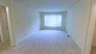 This single-floor space features a generously sized main living area defined by its clean, minimalist design and neutral color palette. The room is finished with wall-to-wall carpet in a light tone, contributing to a soft and comfortable underfoot feel. Smooth, cream-colored walls extend to a standard ceiling height, creating an open and airy atmosphere. A large window dressed with vertical blinds serves as the focal point of the main living area, allowing an abundance of natural light to fill the room while offering a view of the surrounding greenery outdoors. The window also features a baseboard heating unit beneath it, indicating a functional heating system integrated into the design. A floor vent is also visible, suggesting a central air system for year-round climate control. A door positioned along one wall provides access to an adjacent space or room. The overall ambiance of the space is bright, clean, and move-in ready, offering a blank canvas well-suited for a variety of furnishing arrangements and interior styles.