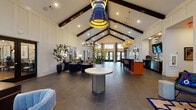 This beautifully designed single-floor community clubhouse or leasing center makes a striking first impression with its soaring vaulted ceilings accented by bold, dark wood beams and a dramatic peaked roofline. The open floor plan is anchored by large-format slate-toned tile flooring that flows throughout, creating a cohesive and expansive feel. A sculptural pendant light in navy and gold serves as a centerpiece overhead, complemented by woven rattan pendants and a circular chandelier that add layers of visual interest.

The main gathering area features a sophisticated lounge with a deep navy tufted sofa, cognac leather armchairs, and a geometric hexagonal wood coffee table, all set atop a richly patterned area rug beneath original abstract artwork. Adjacent seating nooks offer intimate conversation spaces, with floor-to-ceiling floral drapery and French doors that invite natural light and views of an outdoor pool area.

A well-appointed refreshment bar is outfitted with dark cabinetry, a gold-accented faucet, stainless steel under-counter refrigerators, and a bold black-and-white geometric tile backsplash. Curated snacks and beverages are thoughtfully displayed for guests. The white shiplap-style paneled walls throughout the space provide a clean, modern contrast to the eclectic furnishings, artwork, and colorful accents that give this space its distinctive, welcoming character.