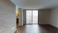 This single-floor residence offers a clean, functional layout with a welcoming open-concept design. The living area is finished with rich, dark hardwood-style flooring that extends throughout, adding warmth and visual continuity to the space. A large sliding glass door fitted with vertical blinds serves as a focal point of the living room, flooding the interior with natural light and providing a view to the outdoors. The white walls and smooth ceiling keep the space feeling bright and airy, while a distinctive exposed brick column adds a touch of texture and character to the design. Adjacent to the living area, the kitchen is accessible through an open passageway, showcasing warm wood cabinetry, stone-look countertops, and dark appliances. The layout encourages a natural flow between the living and kitchen areas, making the space both practical and inviting. Subtle architectural details, including a recessed vent and clean baseboards, reflect a well-maintained interior. Overall, the space strikes a balance between modern simplicity and classic design elements, offering a comfortable and functional living environment suited for everyday life.