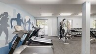 Located at 615 South Kenmore Avenue in Los Angeles, CA, Kenmore Fitness is a dedicated exercise room designed to support a well-rounded workout routine. The space is thoughtfully laid out across a single floor, offering a comfortable and functional environment for physical activity.

The room is immediately distinguished by its eye-catching wall murals depicting athletic silhouettes in motion, branded with "Kenmore Fitness" signage, giving the space an energetic and motivating atmosphere. Light gray carpeting covers the floor throughout, while bright fluorescent lighting ensures the entire area is well-illuminated.

Cardio equipment includes multiple treadmills and an elliptical machine, positioned near a large sliding glass door and window that allow natural light to filter in and provide outdoor views of surrounding greenery. A multi-station cable machine serves as the centerpiece for strength training, complemented by a dedicated dumbbell rack stocked with a range of free weights and a weight bench. Additional accessories such as medicine balls and a stationary bike round out the available equipment.

The room also features two private doors along one wall, a wall-mounted thermostat for climate control, and ample open floor space for stretching or floor exercises. The clean, white walls and well-organized layout contribute to an efficient and welcoming fitness environment.