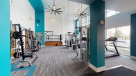 This expansive fitness center is designed to accommodate a wide range of workout disciplines across multiple distinct zones. The space is defined by soaring double-height ceilings in the main strength training area, complemented by bold teal accent walls and white painted brick that together create a vibrant, energizing atmosphere. Oversized pendant lights and large ceiling fans are suspended throughout, adding both functionality and an industrial-chic aesthetic.

The strength training zone is well-equipped with cable machines, a squat rack, barbells, and a comprehensive dumbbell rack, alongside large wall-mounted mirrors that enhance the sense of space. A raised boxing ring with surrounding ropes serves as a standout centerpiece, offering a dedicated combat training area complete with punching bag wall mounts and wooden steps for access.

A separate cardio and cycling studio features floor-to-ceiling windows that invite abundant natural light and outdoor views, housing rowing machines, spin bikes, and a branded rubber flooring design. An entry area provides built-in cubbies, a water station, and a beverage cooler for added convenience. Throughout, textured carpet tile flooring in neutral tones with teal inlay details ties all zones together cohesively.