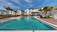 Located at 3050 Millennium Way in Kissimmee, Florida, this expansive outdoor amenity area is designed to deliver a resort-style experience within a residential community. At its centerpiece is a generously sized swimming pool with crystal-clear water, framed by a wide, tile-paved pool deck. Vibrant yellow and navy lounge chairs are arranged throughout the sun deck, offering ample space for relaxation beneath the Florida sunshine. Towering palm trees are thoughtfully positioned throughout the grounds, providing natural shade and a tropical ambiance.

A covered pergola serves as a shaded retreat near the pool, while an outdoor grilling and dining station with tables and chairs caters to social gatherings. Manicured green lawn areas border the pool zone and extend into separate outdoor social spaces, where high-top tables and string lights create an inviting atmosphere for evening use. Well-maintained landscaping with layered shrubs and ornamental grasses lines the pathways and building perimeters, enhancing the lush, polished aesthetic.

Wide concrete walkways connect the various amenity zones, ensuring easy navigation throughout. The single-story clubhouse structure features large glass windows and clean white horizontal siding, providing a bright and welcoming architectural backdrop. The entire outdoor area is enclosed by black metal fencing, offering both security and a defined, cohesive sense of place.