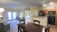 This single-floor residence offers a well-connected, open-concept layout that seamlessly integrates the main living areas. At the heart of the space is a dining area anchored by a dark wood table and matching chairs, positioned adjacent to a raised kitchen counter bar that provides both a functional workspace and a natural divider between the dining and kitchen zones. The kitchen features dark countertops and includes appliances such as a microwave, offering practical convenience.

The living room extends toward a sliding glass door framed by curtains, inviting abundant natural light and providing access to an outdoor area. The space is furnished with comfortable seating, including a blue accent chair, lending a relaxed yet put-together ambiance. Crown molding runs throughout the ceiling, adding an elegant architectural detail against the neutral, warm-toned walls.

Visible from the main living area, the bedroom is accessed through a doorway and features a full-sized bed with crisp white bedding, complemented by warm accent lighting and vibrant green walls that create a lively contrast to the neutral common areas. Carpeting throughout the floors ties the spaces together, contributing to a cozy and cohesive atmosphere across this thoughtfully designed single-level home.