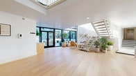 Located at 3650 Regal Pl in the Hollywood Hills neighborhood of Los Angeles, this thoughtfully designed two-story communal space blends modern architecture with an open, airy aesthetic. The main floor is expansive and bathed in natural light, featuring wide-plank light hardwood flooring that runs throughout. Large black-framed floor-to-ceiling glass doors and windows connect the interior seamlessly to the outdoor patio, creating a fluid indoor-outdoor flow. A skylight overhead further amplifies the brightness of the space, lending it a clean, gallery-like quality. A built-in wood reception desk and a dedicated mailroom area anchor the entrance, which also displays curated wall art against crisp white walls. An open-tread white staircase with metal railings leads to the upper level, where neutral carpet, a decorative wall mural, and white railings overlook the main floor below. The outdoor patio features a checkered tile layout, a fire pit lounge area, palm trees, and lush plantings, while a sparkling communal pool sits just beyond, surrounded by tropical landscaping and balconies above. The overall ambiance is bright, modern, and welcoming, with a cohesive neutral palette and abundant natural light throughout both levels.