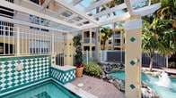 This single-floor residence is part of a vibrant, tropical-inspired multi-story community that immediately impresses with its well-appointed outdoor amenities. The most visually distinctive feature is the communal pool area, which showcases bold green and white checkered tilework along its raised spa or wading section, adding a lively Mediterranean flair to the setting. A white latticed pergola frames the space overhead, filtering sunlight and providing a sense of structure while maintaining an open-air atmosphere. Surrounding the pool, carefully arranged tropical landscaping includes towering palm trees, ornamental shrubs, potted flowering plants, and decorative rock formations, all enclosed by a clean white aluminum fence. The warm yellow exterior of the building complements the vivid tile accents and greenery, reinforcing the resort-style ambiance of the community. Hexagonal paver stones line the pool deck, adding texture and visual interest underfoot. The overall outdoor environment conveys a relaxed, sun-soaked Florida lifestyle. Interior images were not available for review; however, the single-floor layout suggests a straightforward and accessible living arrangement within this amenity-rich setting.