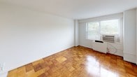 This single-floor studio is characterized by its warm parquet wood flooring, which extends throughout the main living area in a distinctive checkerboard block pattern, adding texture and visual interest to the open layout. The main living space is bright and open, with clean white walls and a window fitted with a window-unit air conditioner that allows natural light and outdoor greenery views to filter in.

The kitchen is efficiently designed and integrated into one end of the unit. It features white cabinetry above and below the counter, a gas range, a single-basin sink, and a compact refrigerator. White ceramic tile flooring delineates the kitchen zone from the parquet of the main living area. A fluorescent light fixture mounted beneath the upper cabinets provides functional task lighting. A shelved closet area is accessible adjacent to the kitchen.

The bathroom is finished with white ceramic tile flooring and retains a classic mid-century character with turquoise accent tiles lining the tub surround. The full tub-shower combination includes grab bars for added accessibility, complemented by a wall-mounted mirror and a vanity sink. Overall, the space presents a clean, functional layout suitable for a range of uses.