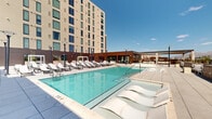 Located in the heart of Tucson, Arizona, this expansive rooftop amenity deck offers a resort-style outdoor experience against a stunning backdrop of the city skyline and the Santa Catalina Mountains. The centerpiece of the space is a large, sparkling swimming pool with in-water lounge chairs and classic depth markings, flanked by rows of white chaise lounges positioned to capture the abundant Arizona sunshine. Adjacent to the pool, a bubbling spa provides a relaxing retreat with open views of the surrounding landscape.

The deck is thoughtfully landscaped with drought-tolerant desert plantings, including agave and native shrubs, integrated into raised masonry planters that reflect the regional aesthetic. Covered pergola structures and shaded seating areas offer respite from the sun, while a covered clubhouse pavilion alongside the pool features a large-format display screen, ceiling fans, and an outdoor mural depicting a vivid Arizona desert sunset — perfect for gatherings and events.

Open-air lounge areas with modern upholstered furniture are scattered throughout the terrace, creating intimate spaces for relaxation while taking in sweeping panoramic views of the Tucson skyline and mountain ranges. The combination of contemporary architecture, desert landscaping, and thoughtfully designed amenities makes this rooftop deck a premier outdoor destination in downtown Tucson.