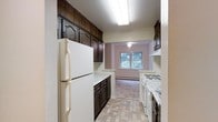 Located at 7 Elk Drive in Monticello, New York, this 2-bedroom, 2-bathroom residence is laid out entirely on a single floor, offering convenient and accessible living throughout. The kitchen is a standout feature, showcasing rich dark wood cabinetry with classic raised-panel detailing that extends to the ceiling, providing generous storage space. Light-toned stone-look countertops offer ample workspace, while white appliances — including a full-size refrigerator and gas range — complete the functional setup. A skylight-style overhead fixture brightens the kitchen, complementing the tile flooring underfoot. The kitchen flows openly into an adjoining room, likely a dining area, which benefits from a window overlooking lush greenery outside, filling the space with natural light and a sense of connection to the outdoors. Soft, neutral wall tones throughout create a calm and welcoming ambiance. A baseboard heating system is visible along the perimeter, ensuring comfortable warmth year-round. The overall layout is practical and well-organized, making efficient use of the available space while maintaining a cozy, residential character suited for everyday living.