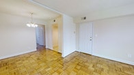 This single-floor space offers a clean, functional layout anchored by distinctive parquet wood flooring that extends throughout the main living area and into the connecting hallway. The warm, honey-toned geometric pattern of the parquet adds visual texture and character to the otherwise neutral interior. Bright white walls and standard-height ceilings create a light, airy atmosphere throughout.

The entry area is framed by a classic six-panel white door with brass hardware, opening directly into the main living space. A partial wall divides the central area, creating a subtle separation between the entry and the interior corridor without fully closing off the flow of movement. This hallway leads further into the unit, with additional rooms accessible through white panel doors, suggesting a practical and connected floor plan.

An elegant chandelier is visible near the hallway entrance, providing a decorative focal point that complements the overall aesthetic. HVAC vents are positioned in the ceiling, ensuring climate control throughout. The combination of warm flooring, white walls, and straightforward architectural elements gives the space a neutral, move-in-ready character well-suited for a variety of uses.