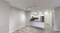 This single-floor residence offers a clean, modern aesthetic defined by an open-concept layout that seamlessly connects the living area to the kitchen. The kitchen is a functional centerpiece, featuring white shaker-style cabinetry, dark granite countertops with a contrasting tile backsplash, and stainless steel appliances including a refrigerator and microwave. A peninsula counter provides a natural division between the kitchen and the living space while offering additional prep and seating space. The living area is bright and airy, with neutral gray walls and crisp white baseboards that complement the warm wood-style plank flooring running throughout the unit. Track lighting in the living area and recessed lighting in the kitchen ensure well-distributed illumination across the open floor plan. The ceilings are smooth and of a comfortable height, contributing to the overall sense of openness. A paneled door visible in the background hints at additional private spaces within the unit. The overall ambiance is contemporary and move-in ready, with a cohesive palette of neutral tones and modern fixtures that create a welcoming, versatile living environment.