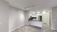 This single-floor residence offers a clean, modern aesthetic defined by an open-concept layout that seamlessly connects the living area to the kitchen. The kitchen is a functional centerpiece, featuring white shaker-style cabinetry, dark granite countertops with a contrasting tile backsplash, and stainless steel appliances including a refrigerator and microwave. A peninsula counter provides a natural division between the kitchen and the living space while offering additional prep and seating space. The living area is bright and airy, with neutral gray walls and crisp white baseboards that complement the warm wood-style plank flooring running throughout the unit. Track lighting in the living area and recessed lighting in the kitchen ensure well-distributed illumination across the open floor plan. The ceilings are smooth and of a comfortable height, contributing to the overall sense of openness. A paneled door visible in the background hints at additional private spaces within the unit. The overall ambiance is contemporary and move-in ready, with a cohesive palette of neutral tones and modern fixtures that create a welcoming, versatile living environment.