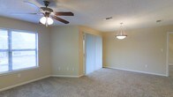 This single-story space presents a clean, functional layout centered around an open living area. The room features wall-to-wall carpeting in a neutral tone, contributing to a warm and comfortable atmosphere. Soft yellow walls provide a welcoming backdrop throughout the space.

Overhead lighting is well-considered, with a multi-bulb ceiling fan positioned in one area of the room to provide both air circulation and ambient light, while a stylish pendant light fixture anchors another section, adding a decorative focal point. The textured ceiling adds subtle visual interest overhead.

A set of sliding glass doors dressed with vertical blinds serves as a prominent feature of the room, allowing natural light to filter in while offering privacy and outdoor access. A window on the adjacent wall further supplements natural lighting, brightening the interior throughout the day.

The room includes standard white baseboards and multiple electrical outlets along the walls, reflecting a practical and straightforward design. The overall ambiance is neutral and move-in ready, offering a versatile canvas suitable for a variety of furnishing arrangements and personal styles.