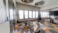 This contemporary fitness center is thoughtfully designed to inspire an active lifestyle. The space centers around a well-equipped cycling studio, where multiple stationary bikes are arranged to take full advantage of the bright, expansive windows that line the walls, flooding the room with natural light. The striking geometric-patterned flooring in bold shades of orange and gray adds a vibrant, energetic character to the space, making it visually dynamic and motivating for workouts. The ceiling features elegant coffered detailing with recessed lighting, adding architectural depth and a refined aesthetic to the room. Full-length mirrors along one wall enhance the sense of openness and allow users to monitor their form during exercise. The neutral gray walls provide a clean backdrop that keeps the focus on the equipment and the bright atmosphere. Decorative tall plants add subtle touches of life and warmth to the environment. The overall ambiance strikes a balance between high-energy functionality and modern design sophistication, creating a fitness environment that is both practical and visually appealing. The single-floor layout ensures easy accessibility throughout the facility.