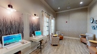This single-floor space functions as a combined lounge and business center, designed with a contemporary aesthetic that emphasizes comfort and functionality. The interior is characterized by light wood-look flooring that extends throughout, contributing to a warm, cohesive atmosphere. Neutral tones dominate the color palette, complemented by textured wave-pattern accent walls that add visual depth and dimension to the space.

The lounge area features multiple patterned armchairs arranged around compact yellow round tables, creating intimate seating clusters ideal for casual conversation or relaxed reading. Wall-mounted magazine racks and a large clock accent one corner, adding a practical and decorative touch. Sheer, scroll-embroidered curtains dress the windows, allowing natural light to filter through while maintaining a soft, refined look.

The business center portion includes wall-mounted white desks with desktop computers, illuminated by industrial-style sconce lighting. Adjustable bar-height stools accompany each workstation, offering a functional yet stylish setup. A wall-mounted display screen is positioned prominently above the workstations.

Recessed ceiling lighting provides consistent illumination throughout, while decorative nature-inspired artwork adorns the walls. An open doorway connects this space to an adjacent area, enhancing the overall sense of openness and flow.