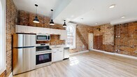 Located at 208 High St in Buffalo, NY, this two-story, 1-bedroom, 2-bathroom unit masterfully combines historic character with contemporary design. The main floor opens to a bright, open-concept living and kitchen area where exposed red brick walls serve as a striking architectural centerpiece. The modern kitchen features white shaker cabinetry, stainless steel appliances including a range, dishwasher, microwave, and refrigerator, complemented by pendant lighting and a large arched window that floods the space with natural light. Light wood flooring runs throughout both levels, enhancing the airy feel of the open layout. A half bathroom on the main floor showcases dramatic dark exposed brick walls paired with a clean white vanity and tile flooring, creating a bold industrial-chic aesthetic. The staircase leads to the upper level, where the spacious primary bedroom features sloped ceilings, additional exposed brick, and an arched window with open views of the surroundings. A dedicated closet and a second full bathroom with a round mirror and white vanity complete the upper floor. The two-story layout, distinctive brickwork, and thoughtful modern updates give this space a unique character that honors its historic Buffalo roots.