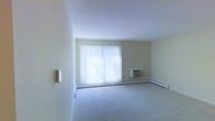This single-floor space offers a clean, functional layout with a focus on simplicity and practicality. The main living area is finished with wall-to-wall carpet in a neutral tone, providing a comfortable and quiet underfoot feel. Light-colored walls contribute to a bright, open atmosphere throughout the room.

A large picture window serves as the focal point of the living area, fitted with vertical blinds that allow for adjustable natural light control. When fully illuminated, the window floods the room with natural light, enhancing the sense of openness in the space. The window reaches close to the floor and is accompanied by a continuous baseboard heating unit that runs along the perimeter of the room, ensuring even heat distribution.

A wall-mounted air conditioning unit is positioned beside the window, providing climate control during warmer months. The ceiling features a smooth, textured finish, and the overall design of the space is practical and understated. A light switch and a small wall-mounted fixture are visible, adding to the functional character of the interior. The space is vacant, allowing its layout and proportions to be clearly appreciated.