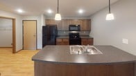 This single-floor residence offers a functional and cohesive layout designed for comfortable everyday living. The kitchen serves as a central hub of the space, featuring warm medium-toned wood cabinetry, dark laminate countertops, and a curved peninsula with a double stainless steel sink. Black appliances, including a refrigerator, range, and built-in microwave, complement the cabinetry, while recessed lighting and a pendant fixture provide focused illumination. The kitchen opens directly to the living area, which is finished with neutral-toned carpet and baseboard heating. A sliding glass door floods the living room with natural light and provides views of lush outdoor greenery, enhancing the sense of openness in the space.

The bedroom is a well-sized, carpeted room with a large window framing outdoor views, baseboard heating, and a closet accessed through a wood-trimmed door. The bathroom is finished with light wood-look flooring, a long vanity with a white countertop and warm wood cabinetry below, a large mirror, and a full tub and shower combination. The overall aesthetic throughout the unit is clean and neutral, with consistent wood-toned accents tying each room together cohesively.