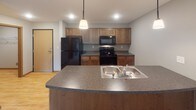 This single-floor residence offers a functional and cohesive layout designed for comfortable everyday living. The kitchen serves as a central hub of the space, featuring warm medium-toned wood cabinetry, dark laminate countertops, and a curved peninsula with a double stainless steel sink. Black appliances, including a refrigerator, range, and built-in microwave, complement the cabinetry, while recessed lighting and a pendant fixture provide focused illumination. The kitchen opens directly to the living area, which is finished with neutral-toned carpet and baseboard heating. A sliding glass door floods the living room with natural light and provides views of lush outdoor greenery, enhancing the sense of openness in the space.

The bedroom is a well-sized, carpeted room with a large window framing outdoor views, baseboard heating, and a closet accessed through a wood-trimmed door. The bathroom is finished with light wood-look flooring, a long vanity with a white countertop and warm wood cabinetry below, a large mirror, and a full tub and shower combination. The overall aesthetic throughout the unit is clean and neutral, with consistent wood-toned accents tying each room together cohesively.