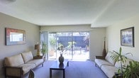 This two-story home offers a well-designed living space with a focus on natural light and indoor-outdoor connectivity. The living room serves as a central gathering area, featuring plush wall-to-wall carpeting in a soft blue-grey tone that adds warmth and comfort underfoot. Neutral-toned walls create a versatile backdrop, while a textured ceiling adds subtle architectural detail.

A prominent sliding glass door spans much of the rear wall, flooding the room with natural light and providing direct access to a private outdoor patio. The patio is enclosed by a wood privacy fence and features outdoor seating, potted plants, and decorative greenery, creating an inviting extension of the interior living space. Lush plantings visible beyond the fence contribute to a serene, garden-like atmosphere.

The living room is generously proportioned, comfortably accommodating multiple seating arrangements, side tables, and a central coffee table. The overall ambiance is calm and understated, with the interplay of natural light and neutral finishes giving the space an open, airy feel. The two-floor layout suggests additional living or sleeping quarters above, offering practical separation between communal and private areas of the home.