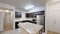 This 2-bedroom, 2-bathroom residence at 15450 SW Scholls Ferry Rd. offers a practical and well-designed single-floor layout. The kitchen serves as a standout feature, outfitted with rich dark wood cabinetry, light granite-style countertops, stainless steel appliances including a refrigerator, range, microwave, and dishwasher, and a central island that provides additional prep and dining space. Light wood-style flooring runs throughout the kitchen, complementing the clean, neutral aesthetic. The open sightline from the kitchen to the living area creates a connected, functional flow. The living area features soft carpeting and a window that frames lush green views. Both bedrooms are carpeted and finished in neutral tones, with the primary bedroom offering a generous footprint and natural light. The two bathrooms are similarly styled, each featuring dark vanity cabinetry, light countertops, tile-patterned flooring, and accessibility grab bars. A dedicated in-unit laundry area accommodates a full-size washer and dryer with overhead shelving. A hallway connects the private quarters of the home, and a closet provides additional storage. The overall interior presents a clean, neutral palette suited for everyday living.