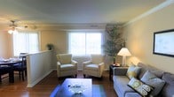 This single-floor space centers around a well-appointed living room that offers a comfortable and inviting atmosphere. The room features warm hardwood flooring that runs throughout, complemented by neutral-toned walls that create a clean, airy backdrop. Crown molding along the ceiling adds a touch of architectural refinement. A large window fitted with vertical blinds serves as the focal point of the room, allowing abundant natural light to fill the space while offering views of the outdoors. The seating arrangement includes two matching cream-colored armchairs positioned symmetrically in front of the window, paired with a gray sofa adorned with patterned accent pillows. A dark-finished coffee table anchors the seating area, styled with a decorative centerpiece. A side table with a lamp provides warm ambient lighting. Lush indoor plants, including a tall palm-style plant in the corner, introduce a refreshing natural element that enhances the room's overall warmth. A ceiling vent ensures comfortable climate control throughout. The overall ambiance is relaxed yet polished, making the space feel both functional and welcoming.