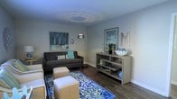 This single-floor space showcases a thoughtfully designed living room that combines comfort and style through a carefully curated aesthetic. The room features dark hardwood flooring that grounds the space and provides a rich contrast to the light gray walls and white ceiling, which is accented by a subtle circular light fixture that adds a decorative touch overhead.

The living area is anchored by a dark charcoal sofa adorned with teal and patterned accent pillows, complemented by a pair of upholstered ottomans that serve as both seating and a functional coffee surface. A bold blue-and-white floral area rug adds a vibrant focal point beneath the seating arrangement, tying the color scheme together cohesively.

A sleek arc floor lamp with a chrome finish provides ambient lighting while adding a modern design element. Along one wall, an open shelving console in a weathered gray finish displays decorative objects, framed artwork, and greenery, contributing to the room's layered, curated feel. Framed wall art in cool blue tones echoes the overall palette, while a large decorative letter serves as an additional personalized accent.