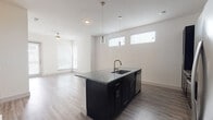 This well-appointed single-floor residence offers a clean, contemporary design with an open-concept layout that seamlessly connects the kitchen, dining, and living areas. The kitchen is a standout feature, showcasing rich dark cabinetry, granite countertops with subtle gray and white veining, a geometric tile backsplash, and a full suite of stainless steel appliances including a refrigerator with ice maker, range, and microwave. A central island with an undermount sink and elegant pendant lighting serves as both a functional workspace and a natural gathering point. Light wood-look flooring flows throughout the main living areas, complementing the neutral white walls and creating a bright, airy atmosphere. The living area benefits from multiple windows fitted with blinds, allowing for natural light control, and is finished with a ceiling fan for year-round comfort. A sliding glass door provides access to an outdoor area. The bedroom features plush carpeting and a large window that fills the room with natural light. The bathroom is equally well-finished, offering a dual-sink vanity with granite countertops, dark cabinetry, a glass-enclosed walk-in shower, and convenient access to a spacious walk-in closet with built-in shelving.