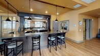 Located in Ramsey, Minnesota, this single-floor community space is thoughtfully designed for socializing and dining. The main dining area features rich hardwood-style flooring, warm golden-orange walls, and a coffered drop ceiling with recessed lighting that creates a comfortable and inviting atmosphere. Multiple square and round dark wood tables are paired with cushioned chairs, offering flexible seating arrangements throughout the open room.

Large windows fitted with horizontal blinds line the perimeter, allowing generous amounts of natural light to flood the space while providing views of the surrounding neighborhood. A central door provides direct access to an outdoor balcony or patio area, extending the usable space beyond the interior.

The bar area is a standout feature, boasting a sleek countertop with bar stool seating, pendant lighting hanging from a dark track system, and a pass-through window connecting to a back-of-house service area equipped with stainless steel fixtures. This setup makes the space well-suited for events and gatherings.

A lounge corner rounds out the space, featuring a wall-mounted flat-screen television, display cabinetry with glass-front doors, and décor accents that add a personalized touch. The overall design balances functionality with warmth, making it a versatile and welcoming venue for community activities.
