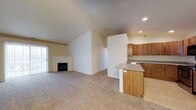 This single-floor space showcases an open-concept layout that seamlessly connects the living area and kitchen, creating a functional and inviting environment. The living room is highlighted by dramatic vaulted ceilings that lend a sense of spaciousness and airiness throughout. A classic fireplace serves as a focal point, adding warmth and character to the main living area. Neutral-toned carpeting in the living space transitions to tile flooring in the kitchen, clearly delineating the two zones while maintaining a cohesive aesthetic. Natural light enters through a sliding glass door or large window on the far wall, brightening the open interior. The kitchen is well-appointed with warm-toned wood upper cabinets, ample counter space, and a built-in sink, all complemented by a peninsula island with light wood paneling that provides additional workspace and potential seating. Recessed lighting in the kitchen ensures the area is well-lit for everyday use. The clean, neutral color palette throughout — from the soft beige walls to the light countertops — creates a versatile backdrop suited to a variety of interior styles. Overall, the space offers a practical and comfortable layout with notable architectural character.
