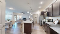 Located on Nichols Loop in Winter Haven, FL, this well-appointed 2-bedroom, 2-bathroom residence offers a thoughtfully designed single-floor layout that maximizes both functionality and comfort. The open-concept living area seamlessly connects the kitchen, dining, and living spaces, creating an inviting atmosphere filled with natural light from multiple windows dressed with stylish curtains. Wood-style plank flooring flows consistently throughout, enhancing the cohesive feel of the home. The kitchen is a standout feature, boasting rich dark cabinetry, granite countertops, a large center island with pendant lighting and bar seating, and a full suite of stainless steel appliances including a refrigerator, range, microwave, and dishwasher. The dining area provides ample space for entertaining, while the living room accommodates comfortable seating and is equipped with a ceiling fan. Both bedrooms are generously sized, each featuring ceiling fans, wood-style flooring, and windows that invite natural light. The two bathrooms include a tub-shower combination with clean white tile. A dedicated laundry room adds everyday convenience. Two closets provide practical storage throughout the home. This residence delivers a modern, functional living experience in a clean and bright environment.