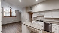 This single-floor unit showcases a thoughtfully designed interior finished with contemporary materials throughout. The kitchen serves as a central highlight, featuring white shaker-style cabinetry, granite-patterned countertops, and a central island that provides additional prep and seating space. Stainless steel appliances, including a refrigerator, range, microwave, and dishwasher, are integrated seamlessly into the layout. A black undermount sink and a stylish multi-tiered pendant light add refined touches. The kitchen benefits from notably elevated ceilings, enhancing the sense of openness in the space.

Wood-look plank flooring extends consistently throughout the unit, providing a cohesive and durable foundation. The living and bedroom areas are generously proportioned, featuring neutral gray walls, clean baseboards, and ample electrical outlets positioned for flexible furniture arrangements. A bedroom area includes a wood-framed window with distinctive gridded panes, introducing natural light and architectural character.

The bathroom is functional and well-organized, with a walk-in shower unit, toilet, vanity with storage cabinetry, and warm-toned wood-look flooring. Overall, the unit presents a clean, modern aesthetic with practical finishes suited for everyday living.