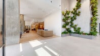 This modern lobby makes an immediate impression with its soaring ceiling heights, crisp white walls, and large-format light stone tile flooring that seamlessly transitions to pale hardwood throughout the interior zones. A dramatic living green wall — cascading with lush, varied plantings arranged across a textured concrete surface — serves as the space's defining centerpiece, bringing a vibrant, organic element to the otherwise clean, minimal aesthetic.

Natural light pours in through expansive floor-to-ceiling steel-framed windows, offering street views and filling the space with warmth throughout the day. A sleek, wood-paneled reception desk anchors the entry, providing a welcoming focal point upon arrival. Exposed concrete columns punctuate the open plan, lending an industrial counterpoint to the refined finishes.

Distinct lounge areas are thoughtfully arranged throughout, featuring contemporary upholstered seating, marble-patterned area rugs, and curated open shelving with decorative objects. A glass-enclosed private room with black steel framing offers a quiet retreat within the larger space. The dedicated mailroom is outfitted with extensive built-in mailboxes framed by warm wood cabinetry and illuminated by sleek linear ceiling lighting. Together, these elements create a cohesive, design-forward environment that balances warmth, functionality, and architectural distinction.