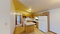 This single-floor, 2-bedroom, 1-bathroom residence offers a practical and comfortable layout designed for everyday living. The entrance opens into a spacious living area featuring wall-to-wall carpet, vaulted ceilings, a ceiling light fixture, and a sliding glass door that leads to a private balcony, flooding the space with natural light. The kitchen is well-appointed with warm oak cabinetry offering generous storage, light-toned countertops, a double-basin sink, a full suite of white appliances including a dishwasher, electric range, and refrigerator, and a central island that provides additional prep space. Tile flooring runs throughout the kitchen and connects seamlessly to the hallway. The primary bedroom is generously sized and features soft carpet, vaulted ceilings, a mirrored sliding closet door, and built-in wire shelving for organized storage. The second bedroom is bright and carpeted, with clean white walls and warm wood baseboards. The full bathroom includes a wide vanity with oak cabinetry, a large mirror with overhead lighting, tile flooring, and a baseboard heater. A dedicated closet with wire shelving adds further storage utility. Throughout the home, warm-toned wood trim provides a cohesive and inviting aesthetic.