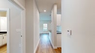 This single-floor unit offers a thoughtfully organized layout comprising a combined kitchen and living room, one bedroom, a dedicated laundry room, and a connecting hallway. The kitchen is a standout feature, showcasing white shaker-style cabinetry with matte black hardware, dark stone countertops, stainless steel appliances including a refrigerator, range, microwave, and dishwasher, and a deep undermount sink. Stylish pendant lights hang above the extended kitchen counter, which also functions as a breakfast bar. The kitchen flows seamlessly into the living area, which benefits from natural light through large windows and a glass door that provides outdoor views and easy access. Wide-plank wood-style flooring runs throughout the main living areas and hallway, lending warmth and continuity to the space. The bedroom is generously proportioned, featuring plush carpet flooring, neutral walls, and a window with outdoor views, creating a calm and comfortable retreat. A dedicated laundry room provides added convenience with wire shelving for storage. The hallway connects all areas efficiently, maintaining a clean, open feel throughout. The overall aesthetic is modern and neutral, with a bright, airy ambiance that makes the space feel welcoming and functional.