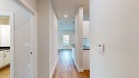 This single-floor unit offers a thoughtfully organized layout comprising a combined kitchen and living room, one bedroom, a dedicated laundry room, and a connecting hallway. The kitchen is a standout feature, showcasing white shaker-style cabinetry with matte black hardware, dark stone countertops, stainless steel appliances including a refrigerator, range, microwave, and dishwasher, and a deep undermount sink. Stylish pendant lights hang above the extended kitchen counter, which also functions as a breakfast bar. The kitchen flows seamlessly into the living area, which benefits from natural light through large windows and a glass door that provides outdoor views and easy access. Wide-plank wood-style flooring runs throughout the main living areas and hallway, lending warmth and continuity to the space. The bedroom is generously proportioned, featuring plush carpet flooring, neutral walls, and a window with outdoor views, creating a calm and comfortable retreat. A dedicated laundry room provides added convenience with wire shelving for storage. The hallway connects all areas efficiently, maintaining a clean, open feel throughout. The overall aesthetic is modern and neutral, with a bright, airy ambiance that makes the space feel welcoming and functional.