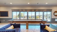 This well-appointed community lounge and game room in Vallejo, California, is designed for social gathering and recreation. The space is defined by a bold two-tone color scheme, with rich teal wainscoting lining the lower walls and crisp white above, creating a sophisticated yet playful atmosphere throughout. Light-toned parquet-style flooring adds warmth and texture underfoot.

At the heart of the room, a full-size pool table with a blue felt surface serves as a central focal point, complemented by a wooden shuffleboard table positioned in front of a sweeping wall of floor-to-ceiling windows. These expansive windows flood the space with abundant natural light and frame panoramic views of the surrounding Vallejo hillside and cityscape beyond.

Plush, channeled blue velvet sofas and accent chairs are thoughtfully arranged throughout, offering comfortable seating for relaxation or conversation. A foosball table adds to the recreational offerings, while curated wall art, including a gallery-style arrangement of framed prints and a vibrant fire-themed display piece, contribute to the room's dynamic, curated aesthetic.

Recessed lighting and pendant fixtures provide layered illumination, while a sliding panel door connects this space to an adjacent dining or meeting area, enhancing the room's versatility. Overall, this lounge delivers an inviting, upscale environment ideal for recreation and community engagement.