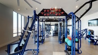 This Dallas fitness center is a comprehensively outfitted training facility designed to meet a wide range of workout needs. The space is anchored by a prominent Hoist multi-station functional training rig finished in bold blue, forming a central corridor of exercise stations loaded with weight plates and cable attachments. The layout is thoughtfully organized, allowing users to move freely between equipment zones without congestion.

The flooring is a durable, low-maintenance surface suited for heavy equipment and high-traffic use. Large windows along the perimeter allow natural light to fill the space, contributing to an open, energizing atmosphere. Recessed ceiling lighting supplements the daylight, ensuring consistent illumination throughout the facility.

A well-stocked dumbbell rack lines one side of the room, offering a full range of free weights for strength training. Medicine balls and additional functional training accessories are readily accessible. Full-length mirrors are positioned strategically to assist with form and technique. The overall design is clean and modern, with white walls and high ceilings enhancing the sense of spaciousness, making this a professional-caliber training environment.