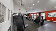Located at 6101 Westgate Dr in Orlando, FL, this fitness center offers a generously sized, single-floor layout designed to accommodate a comprehensive workout experience. The space is defined by its soaring vaulted ceilings, which lend an open and airy atmosphere throughout. Ceiling fans suspended from above provide consistent air circulation, complementing the track lighting that illuminates the workout floor.

The main workout area features durable rubber flooring throughout and is divided into distinct functional zones. A strength training section houses multiple Nautilus and cable resistance machines, adjustable benches, and weight stations, catering to a full range of strength exercises. Cardio equipment, including treadmills and an upright cycle, lines the boldly accented red accent wall, which energizes the space visually. A dedicated stretching and open-floor zone is equipped with stability balls and is flanked by large wall-mounted mirrors that enhance the sense of depth and allow users to monitor their form.

Natural light filters in through several windows fitted with blinds, and a wall-mounted television provides entertainment during workouts. The facility also includes clearly marked emergency exits and informational postings. The combination of high ceilings, vibrant design accents, and a comprehensive equipment selection makes this fitness center a functional and motivating environment for users of all fitness levels.