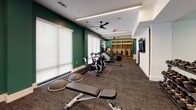 Located at 615 Montgomery St in Savannah, GA, this fitness center offers a well-equipped, single-floor space designed for a comprehensive workout experience. The interior is defined by bold teal-green accent walls contrasted against crisp white trim and ceilings, creating an energetic yet clean aesthetic throughout. Linear LED lighting runs along the ceiling, supplemented by ceiling fans that help maintain airflow and comfort during workouts.

The cardio zone is a standout feature, housing Schwinn spin bikes with integrated monitors, a Concept2 rowing machine, and a Matrix stair climber, all positioned along one side of the room atop protective floor mats. Large roller shade windows line the walls, allowing diffused natural light to fill the space while maintaining privacy.

A built-in wood shelving unit at the far end of the room provides organized storage for accessories such as resistance bands, yoga blocks, and stability balls, keeping the space tidy and functional. A wall-mounted flat-screen TV is positioned for visibility across the main exercise area.

An open floor section accommodates free-form training, with a weight bench, BOSU balance trainers, and a medicine ball rack available for strength and functional fitness work. A secondary door provides additional access. The overall layout is thoughtfully arranged to support a variety of workout styles in a modern, well-maintained environment.