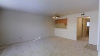 This single-floor residence offers a straightforward, functional layout designed to maximize usability. The living area is generously proportioned, finished with wall-to-wall carpeting in a neutral tone that lends warmth and comfort underfoot. Smooth, white walls and a standard flat ceiling create a clean, bright backdrop throughout the space. A ceiling fan with integrated lighting is centrally positioned, providing both air circulation and ambient illumination to the room.

A defining architectural feature is the pass-through opening connecting the living area to the kitchen, topped with warm wood-toned upper cabinetry. This open-concept design element encourages easy interaction between the two spaces while maintaining a sense of defined zones. The kitchen cabinetry features a classic honey-toned wood finish, adding a touch of warmth to the otherwise neutral palette.

The overall ambiance is calm and understated, making it well-suited for personalization. The neutral color scheme throughout — from the carpeting to the walls — provides a versatile canvas for a variety of interior styles. The space is practical and efficiently organized, with the single-floor plan ensuring seamless flow and easy accessibility between all areas.