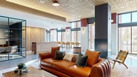 Located at 3720 N Downing St in Denver's vibrant Cole neighborhood, this generously sized lounge and mail room is a thoughtfully designed communal amenity space spread across a single floor. The lounge area immediately impresses with soaring floor-to-ceiling windows that flood the interior with natural light and offer views of the surrounding Denver streetscape. A striking tiled fireplace flanked by bold teal built-in shelving serves as the focal point of the seating area, which features a rich cognac leather sofa, eclectic accent chairs, and layered area rugs atop polished concrete floors. A large-scale colorful mural adds an artistic energy near the entry corridor, complementing the space's contemporary design palette of warm wood tones, deep teal, and terracotta red accents. Textured wood-panel ceilings lend warmth overhead throughout the space. A café-style seating area with bar chairs and large windows offers an additional gathering spot, while a vintage-inspired wood credenza anchors the transitional hallway. The dedicated mail room provides practical functionality with extensive rows of individual resident mailboxes lining the teal beadboard-accented walls, centered around a stone-topped island. Globe sconce lighting and recessed ceiling fixtures ensure the space is well-lit and welcoming.