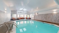 This single-level indoor pool facility is thoughtfully designed to blend recreational function with a refined, resort-like atmosphere. The centerpiece is a generously sized heated swimming pool with crystal-clear turquoise water, illuminated by underwater lighting that enhances the inviting ambiance. The pool is surrounded by a tiled deck that provides ample circulation space.

The architecture features soaring vaulted ceilings with strategically placed skylights that draw in abundant natural light, creating a bright and airy environment. A combination of wall sconces and recessed lighting supplements the natural illumination. The walls are clad in stacked stone tile in cool grey tones, adding a textural, upscale quality to the space, while separate sections feature large-format grey stone-look tile for a clean, contemporary finish.

Multiple seating areas are arranged along the perimeter, furnished with all-weather wicker sofas, lounge chairs, and ottomans dressed in bold orange cushions and accent pillows, offering a warm contrast to the neutral stone surroundings. Large windows throughout the lounge areas frame views of lush greenery outside. A dedicated entry door with safety signage and a life ring station ensure both accessibility and safety. The overall ambiance is polished, comfortable, and well-suited for year-round use.