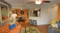 This single-floor residence showcases an open-concept layout that effortlessly connects the living, dining, and kitchen areas into one cohesive and functional space. Rich wood-style flooring runs throughout, anchoring the warm and inviting ambiance. The living room is bright and welcoming, accented by a ceiling fan with a light fixture and crown molding that adds a refined architectural touch. A glass-top coffee table and colorful area rug introduce character and style to the space. The dining area is defined by a bold accent wall in a warm amber tone, complemented by a decorative sunburst wall piece and a round dining table with upholstered chairs — creating an intimate setting for meals. The kitchen is accessible via a peninsula-style breakfast bar with bar stool seating, making it ideal for casual dining and entertaining. The kitchen features dark cabinetry, a refrigerator, and overhead track lighting that brightens the workspace. A pass-through design between the kitchen and living areas enhances the sense of openness. Crown molding, neutral wall tones, and consistent flooring throughout lend the space a polished, cohesive aesthetic that is both practical and visually appealing.