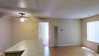 This single-floor space offers an open and functional layout that connects the kitchen and living areas seamlessly. The flooring throughout is hardwood, lending warmth and continuity to the space. A textured popcorn ceiling adds a classic touch overhead, while a ceiling fan with a light fixture provides both airflow and ambient lighting to the main living area.

A tiled countertop is visible in the foreground, marking the transition between the kitchen and the living space. The neutral beige walls create a clean, understated backdrop throughout. A wall-mounted heater unit is positioned along one wall, serving as the primary heating source for the space. Light switch plates and electrical outlets are conveniently distributed along the walls.

A doorway leads to an adjoining room, allowing natural light to filter through and contributing to the overall brightness of the interior. The open sightlines between areas enhance the sense of space, making the layout feel expansive and easy to navigate. Overall, the space presents a straightforward, practical design with durable finishes well-suited for everyday living.