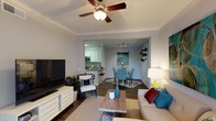 This single-floor residence showcases an open-concept design that thoughtfully connects the living, dining, and kitchen areas into one cohesive and functional space. The living room is anchored by a ceiling fan with a light fixture and elegant crown moulding, lending a polished, finished look to the space. Hardwood-style flooring flows throughout, adding warmth and visual continuity across the main areas.

The living area provides ample room for relaxed seating, complemented by a flat-screen television mounted on a white media console and a stylish accent chair. A wooden coffee table serves as a central focal point, decorated with coastal-inspired accessories in teal and white tones.

Adjacent to the living room, the dining area features a set of teal upholstered chairs around a dark dining table, positioned beneath track lighting that highlights the space. Artwork on the walls reinforces a coastal aesthetic throughout.

The kitchen, visible through an open pass-through, offers white cabinetry, stainless steel appliances, and a breakfast bar with seating, making it both practical and inviting. Overall, the space maintains a bright, airy, and cohesive ambiance.